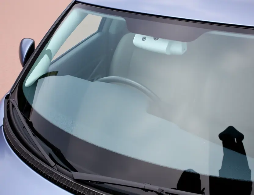 A close-up shot of a Suzuki Swift with a replaced windshield. The windshield is clear and new, contrasting with the older, slightly damaged windshield. The car is parked on a clean, paved surface. Focus on the windshield replacement detail.