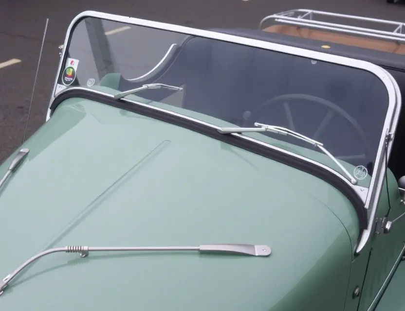 A close-up shot of a Morgan Plus Four car with a replaced windshield. The windshield is clear and new, seamlessly integrated with the existing frame. The car is parked on a clean, slightly reflective surface. Focus on the detail of the new windshield and the car's classic design.