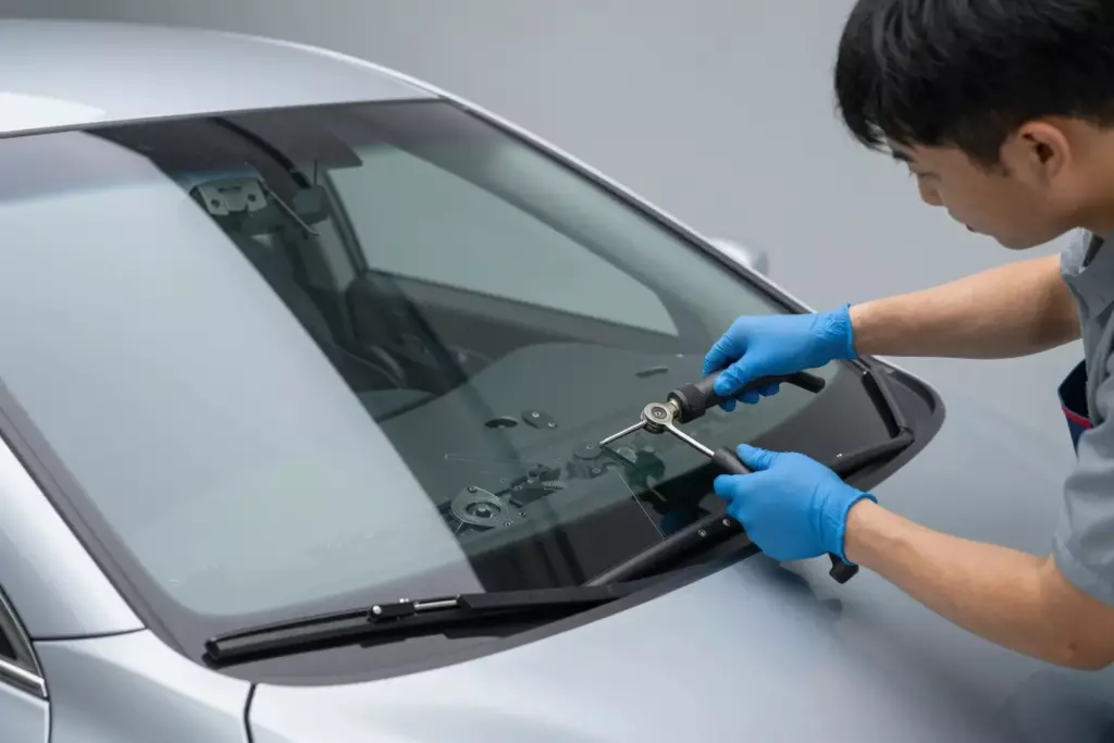 A professional mechanic in a clean, modern workshop wearing blue gloves carefully installing a new windshield into a silver Acura sedan. The car is parked indoors, and the mechanic is using specialized tools to fit the glass precisely. High quality, detailed, realistic lighting.