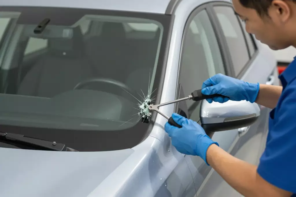 A professional mechanic in a workshop wearing blue gloves carefully removing a cracked windshield from a silver SUV using specialized tools, the car is parked indoors with bright lighting, focus on the hands and the glass edge