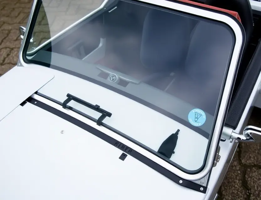 A close-up shot of a Caterham Seven 310 with a replaced windshield. The windshield is clear and new, contrasting with the older, slightly damaged windshield that has been removed. The car is parked on a clean, paved surface. Focus on the details of the replacement process and the quality of the new windshield.